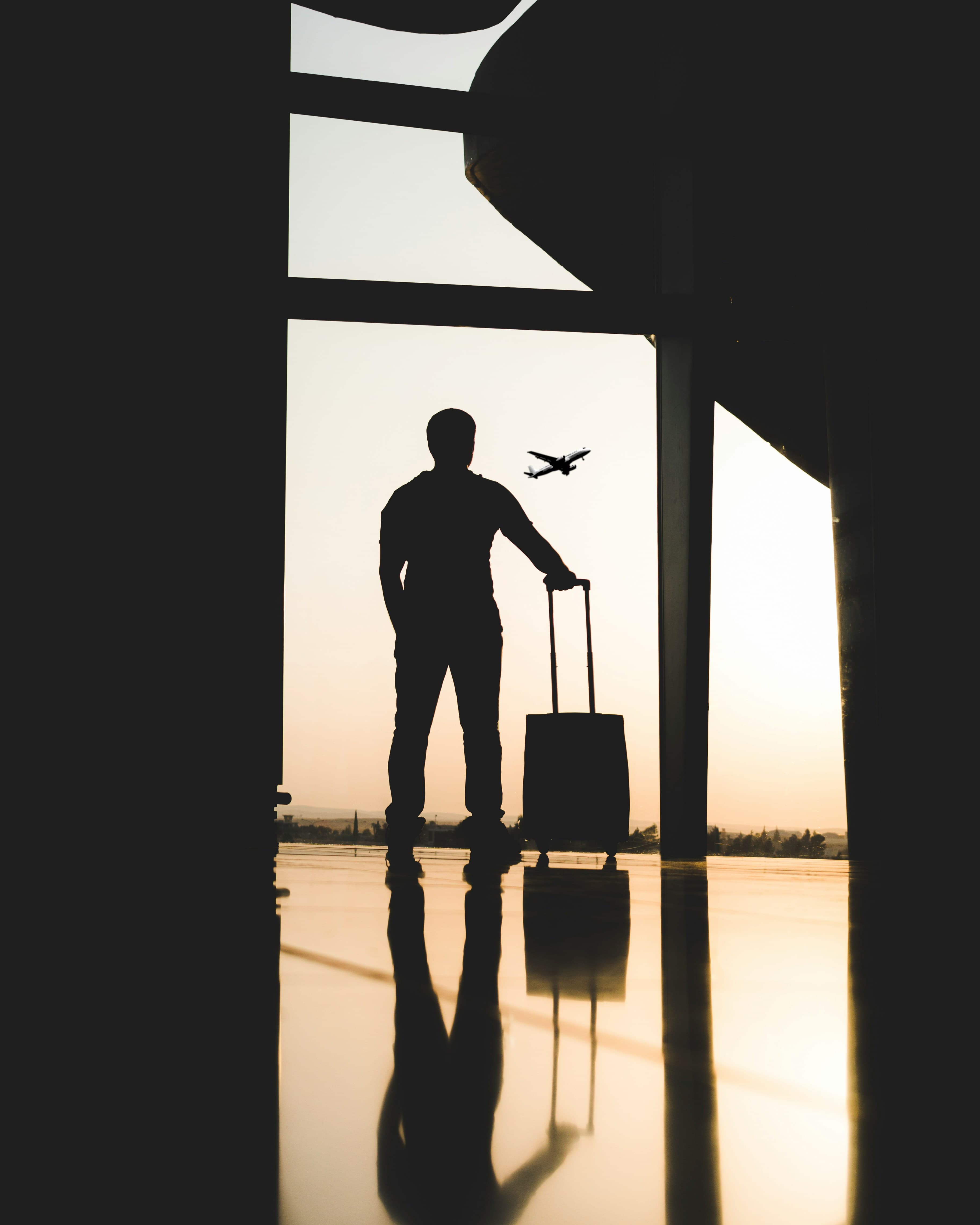 A traveller silhouetted against an airport window at sunset, suitcase in hand, watching a plane take off.